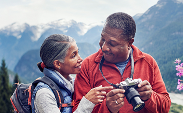 With wooded mountains in the distance, a couple smiles at each
                      other while looking at photos on a camera.