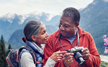 With wooded mountains in the distance, a couple smiles at each
                      other while looking at photos on a camera.